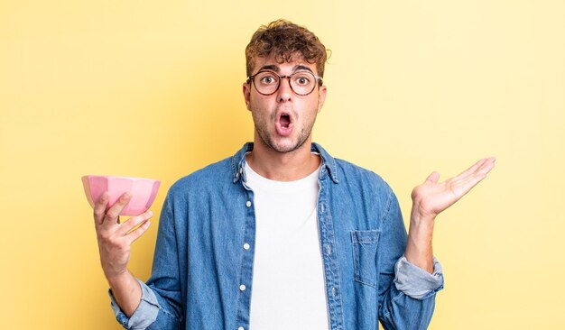 Young handsome man looking surprised and shocked, with jaw dropped holding an object. empty bowl concept