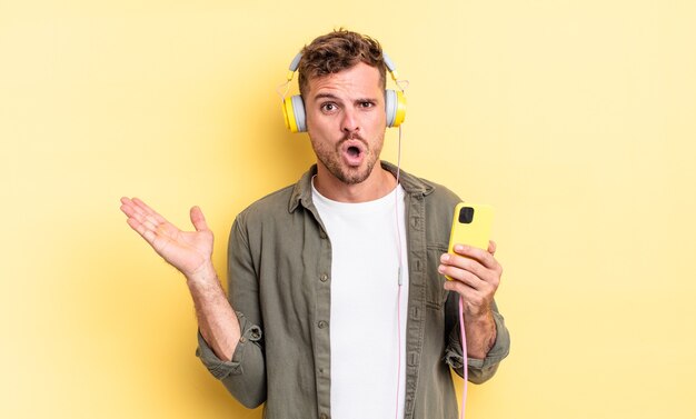 Young handsome man looking surprised and shocked, with jaw dropped holding an object headphones and smartphone concept