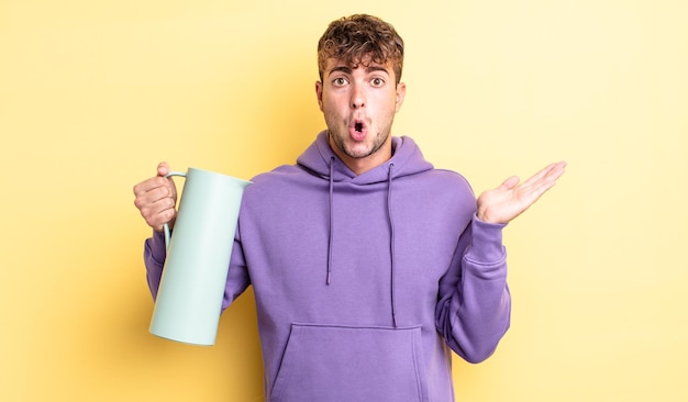 Young handsome man looking surprised and shocked, with jaw dropped holding an object. thermos concept