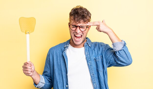 Young handsome man looking unhappy and stressed, suicide gesture making gun sign. flyswatter concept