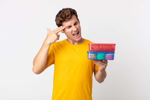 Young handsome man looking unhappy and stressed, suicide gesture making gun sign and holding lunch boxes