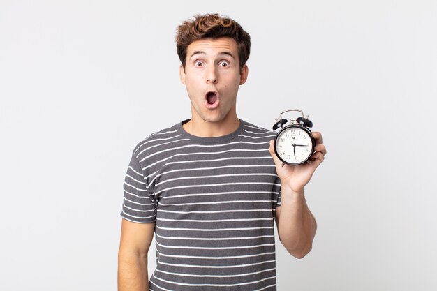 Young handsome man looking very shocked or surprised and holding an alarm clock