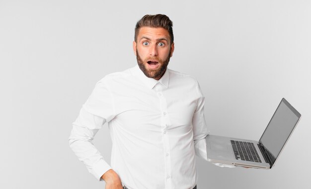 Young handsome man looking very shocked or surprised and holding a laptop