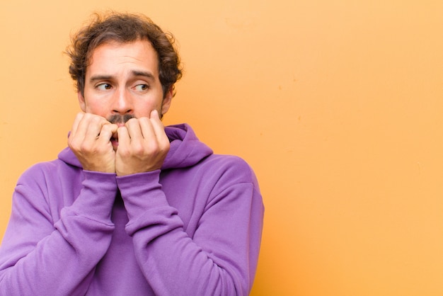 Young handsome man looking worried, anxious, stressed and afraid, biting fingernails and looking to lateral copy space on orange wall