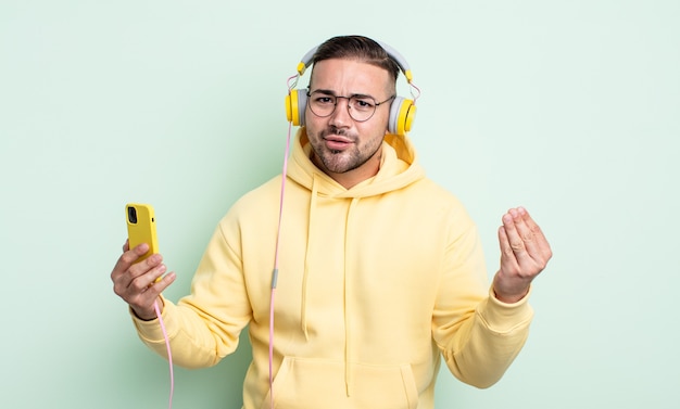 Young handsome man making capice or money gesture, telling you to pay. headphones and smartphone concept