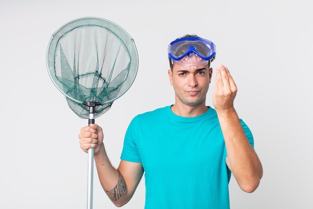 young handsome man making capice or money gesture, telling you to pay with goggles and fishing net