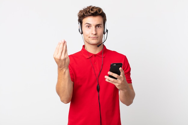 Young handsome man making capice or money gesture, telling you to pay with a smartphone and headset