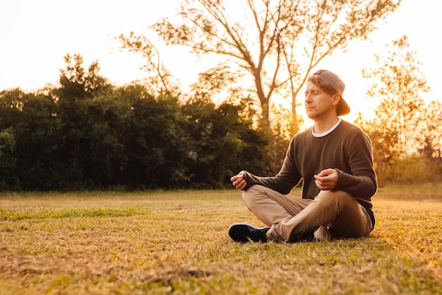 Giovane bell'uomo in posizione meditativa si siede su un prato sullo sfondo della foresta e del tramonto