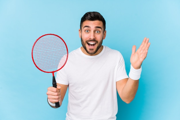 Giovane uomo bello che gioca a badminton isolato ricevendo una piacevole sorpresa, eccitato e alzando le mani.