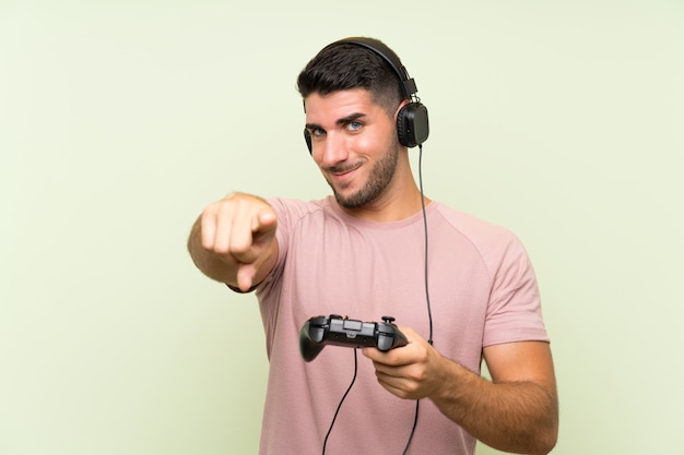 Young handsome man playing with a video game controller over green wall points finger at you with a confident expression