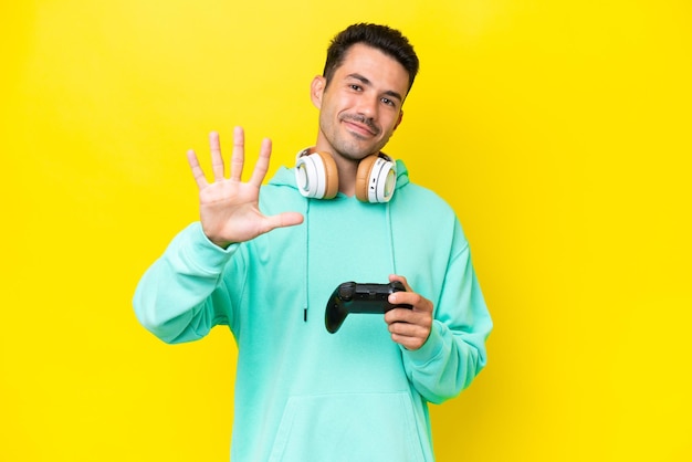 Молодой красивый мужчина играет с контроллером видеоигры над изолированной стеной, считая пять пальцами