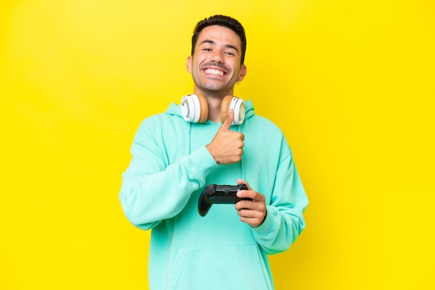 Photo young handsome man playing with a video game controller over isolated wall giving a thumbs up gesture