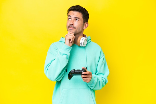 Young handsome man playing with a video game controller over isolated wall having doubts and thinking