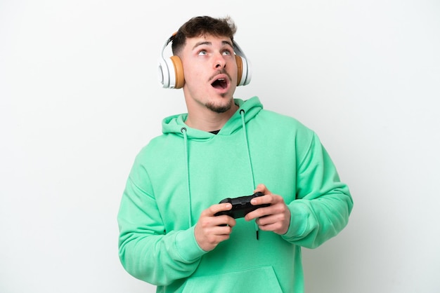 Giovane uomo bello che gioca con un controller per videogiochi isolato su sfondo bianco guardando in alto e con espressione sorpresa