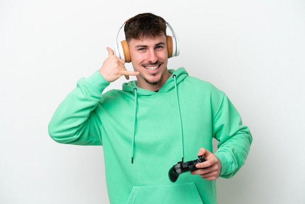 Young handsome man playing with a video game controller isolated on white background making phone gesture Call me back sign