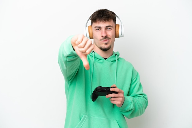 Giovane uomo bello che gioca con un controller per videogiochi isolato su sfondo bianco che mostra il pollice verso il basso con un'espressione negativa