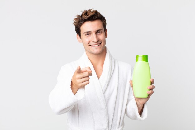 young handsome man pointing at camera choosing you with bathrobe and holding a shampoo bottle