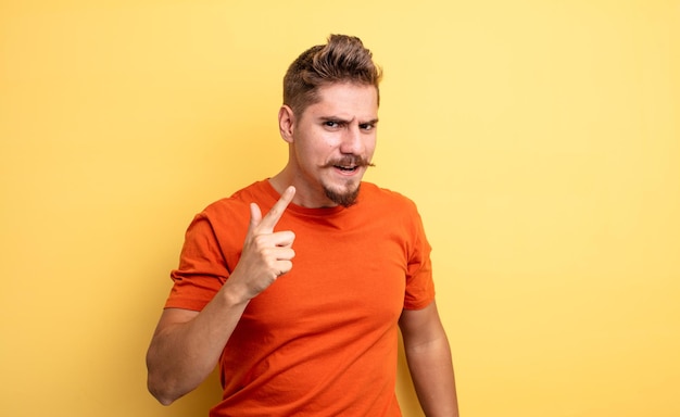 Young handsome man pointing at camera with an angry aggressive expression looking like a furious crazy boss strange moustache concept