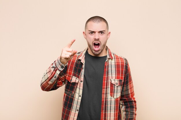 Young handsome man pointing with an angry aggressive expression looking like a furious, crazy boss against flat wall