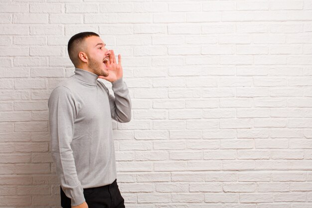 Young handsome man profile view, looking happy and excited, shouting and calling to copy space on the side against flat background