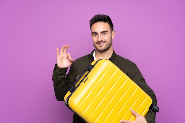 Young handsome man over purple in vacation with travel suitcase and making OK sign