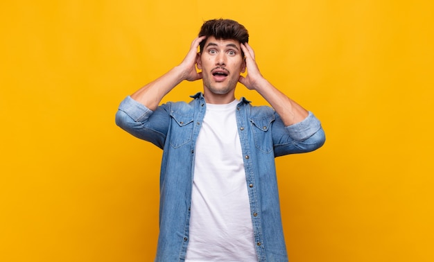 Young handsome man raising hands to head, open-mouthed, feeling extremely lucky, surprised, excited and happy