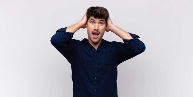 Young handsome man raising hands to head, open-mouthed, feeling extremely lucky, surprised, excited and happy