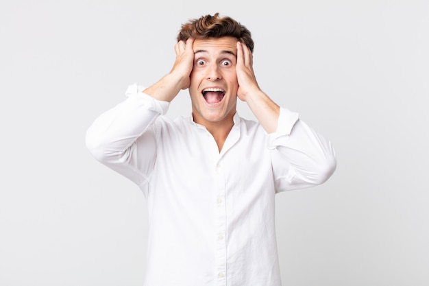 Young handsome man raising hands to head, open-mouthed, feeling extremely lucky, surprised, excited and happy