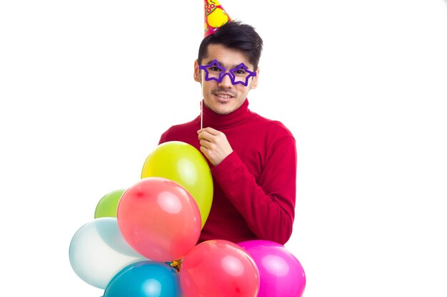 Young handsome man in red shirt with celebrating hat holding many colored balloons and card stick of glasses on white background in studio