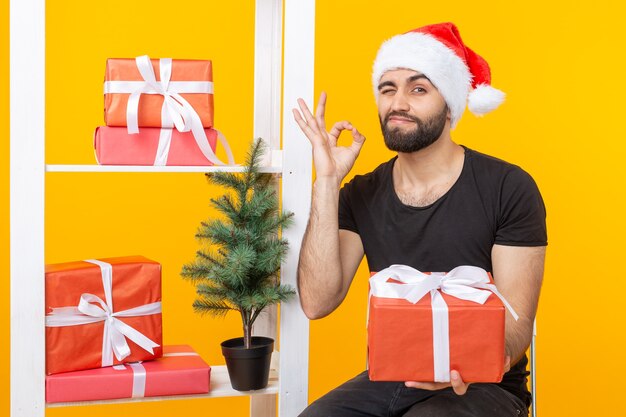 Young handsome man in a Santa Claus hat is holding congratulatory gifts next to a Christmas tree. Concept of holidays of Christmas