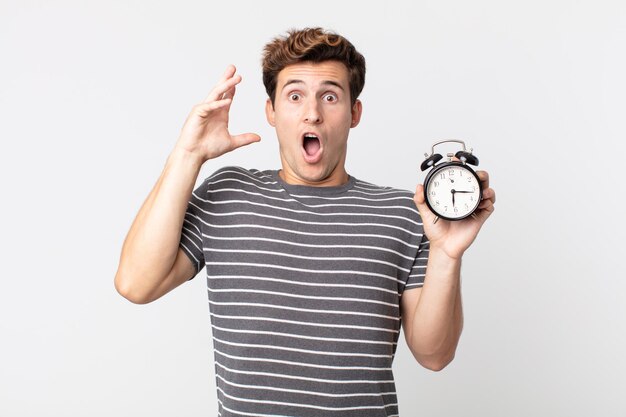 Young handsome man screaming with hands up in the air and holding an alarm clock