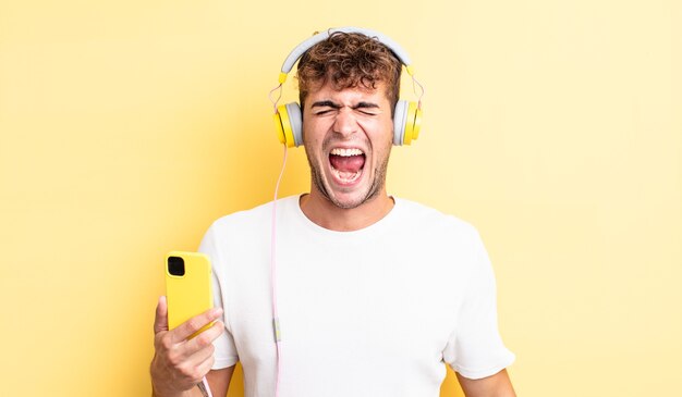 Young handsome man shouting aggressively, looking very angry. headphones and smartphone concept