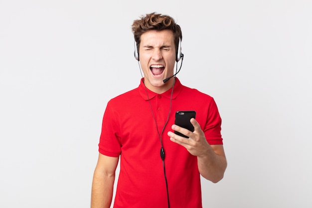 Young handsome man shouting aggressively, looking very angry with a smartphone and headset