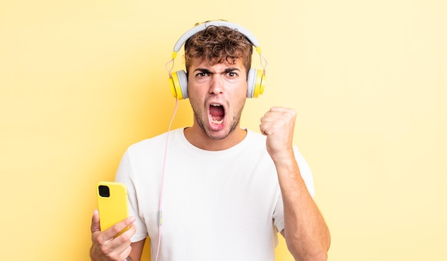 Young handsome man shouting aggressively with an angry expression. headphones and smartphone concept