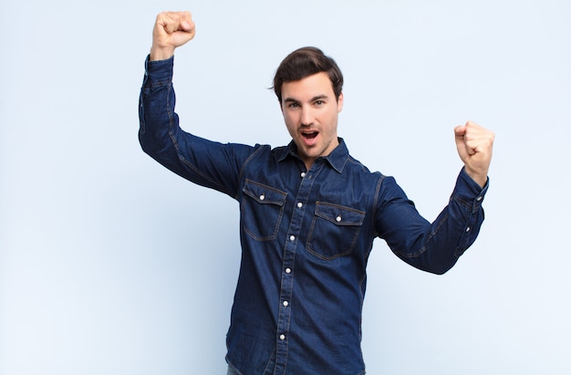 Young handsome man shouting triumphantly, looking like excited, happy and surprised winner, celebrating against blue wall