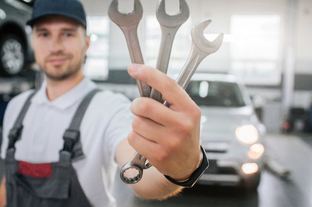 Young handsome man shows set of wrenches. He looks at them and smile. Guy wears uniform. He stands in front of white car. Young man smiles a bit.