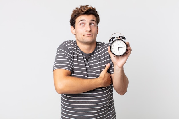 Young handsome man shrugging, feeling confused and uncertain and holding an alarm clock