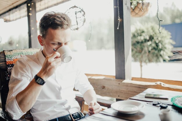 若いハンサムな男はテラスで白いシャツに座ってコーヒーを飲みます