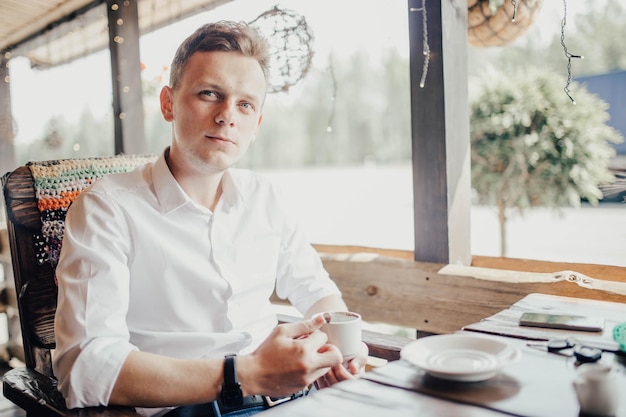 young handsome man sits in a white shirt on the terrace and drinks coffee