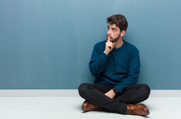 Young handsome man sitting on floor asking for silence and quiet, gesturing with finger in front of mouth