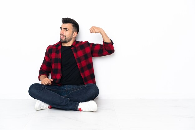 Young handsome man sitting on the floor doing strong gesture