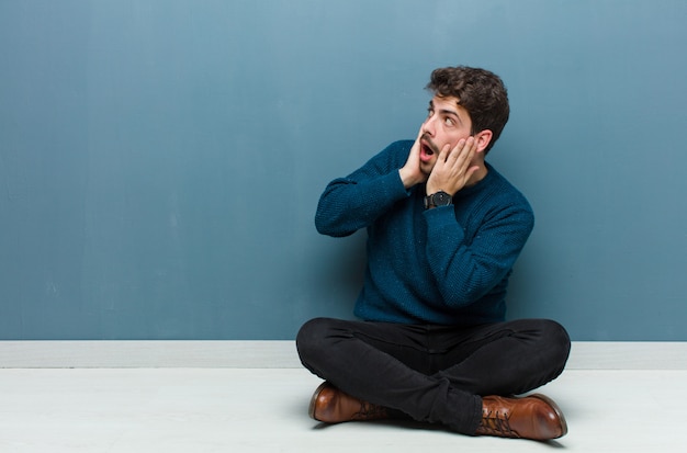 Young handsome man sitting on floor feeling happy, excited and surprised, looking to the side with both hands on face