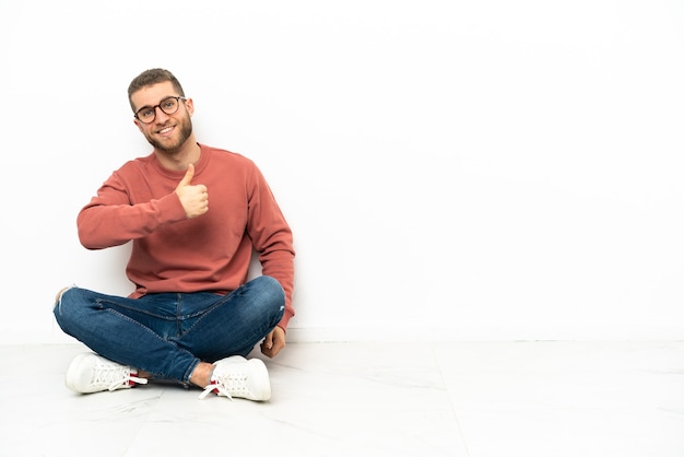 Young handsome man sitting on the floor giving a thumbs up gesture
