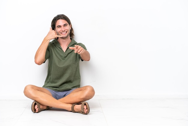 Young handsome man sitting on the floor isolated on white background making phone gesture and pointing front