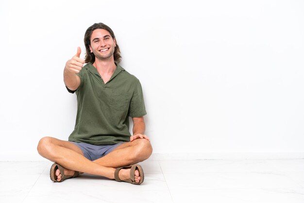 Young handsome man sitting on the floor isolated on white background with thumbs up because something good has happened