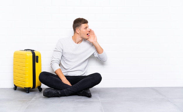 Young handsome man sitting on the floor with a suitcase shouting with mouth wide open