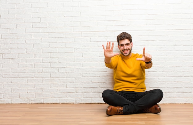 Young handsome man sitting on home floor against brick wall texture