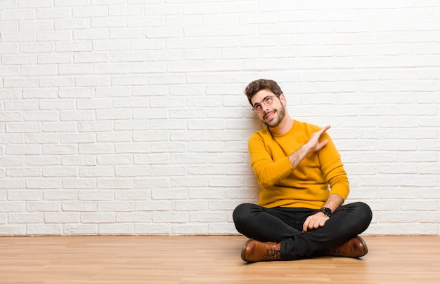 Young handsome man sitting on home floor against brick wall texture