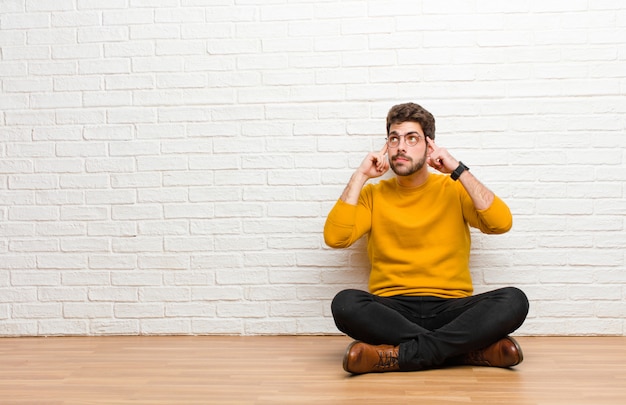 Young handsome man sitting on home floor against brick wall texture