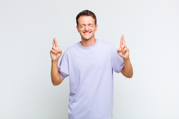 Young handsome man smiling and anxiously crossing both fingers, feeling worried and wishing or hoping for good luck